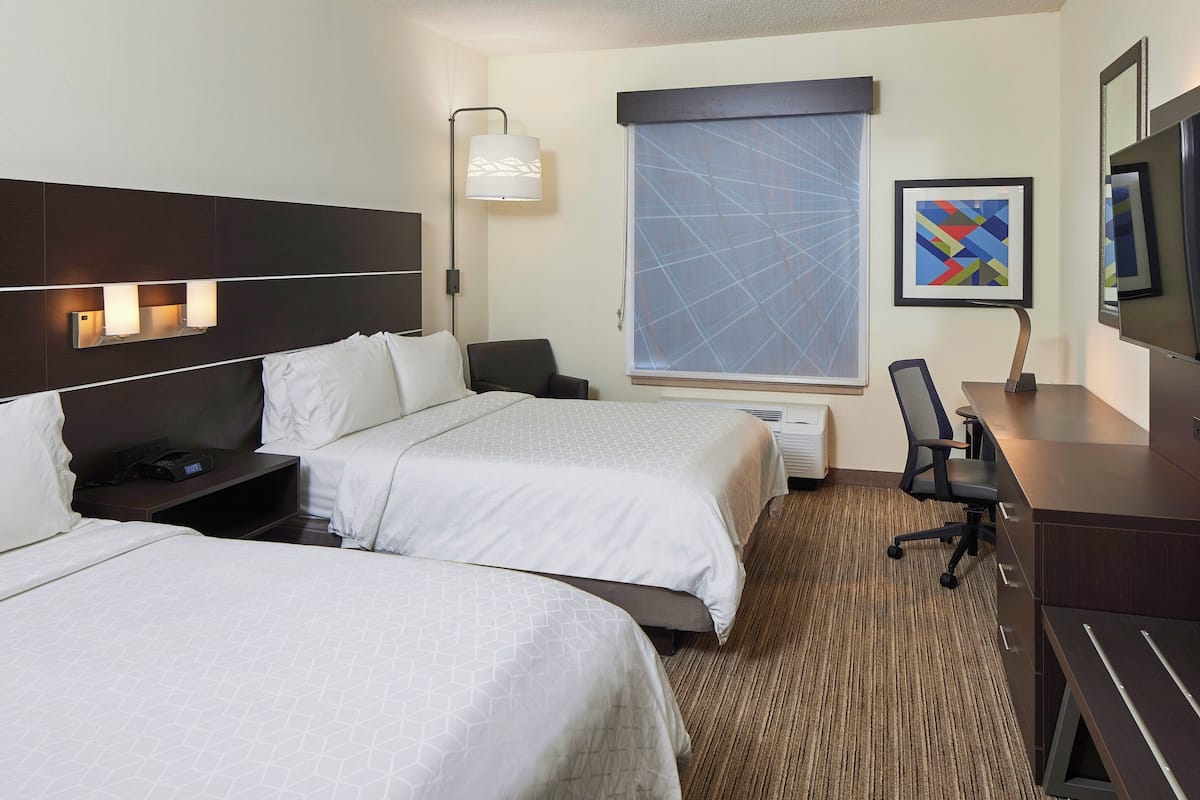 A hotel room with two beds, a desk, a chair, a TV, and a window with blinds.