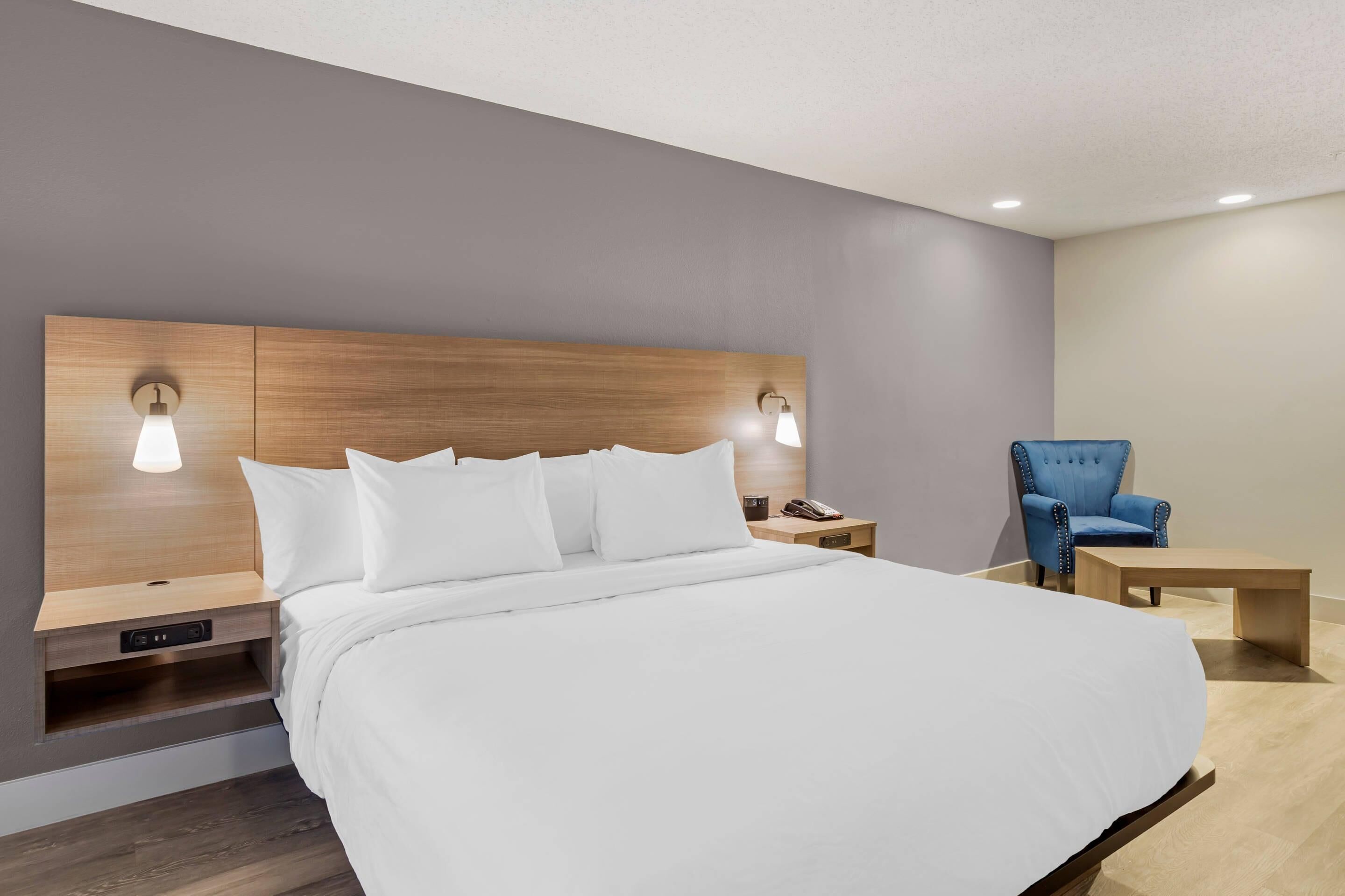 A modern hotel room with a large bed, bedside tables, a blue armchair, and a small wooden table.