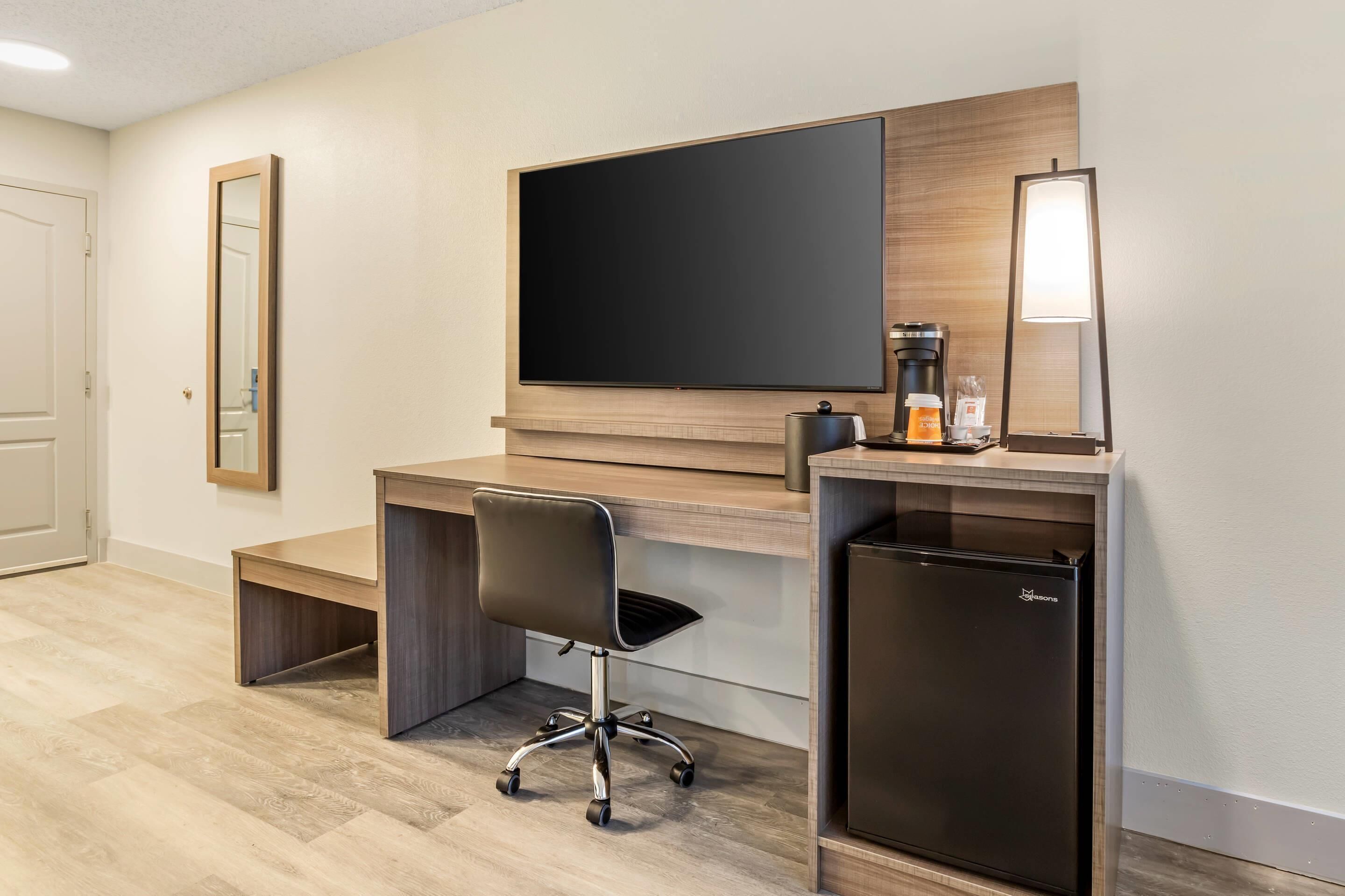 A modern hotel room with a wooden desk, a black office chair, a flat-screen TV, a coffee machine, a mini fridge, and a mirror.