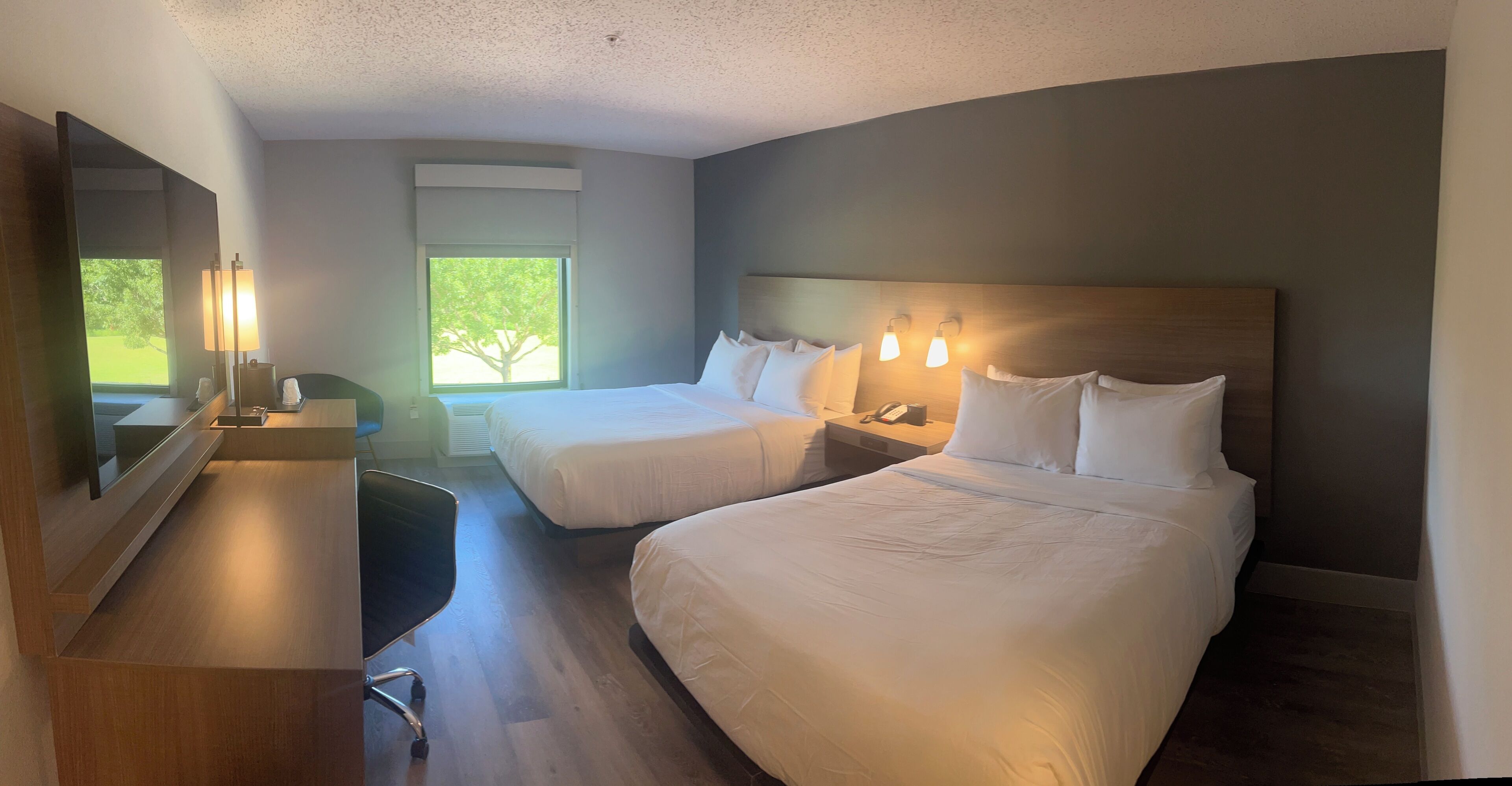 A hotel room with two beds, a desk, a chair, a mirror, and a window with a view of trees.