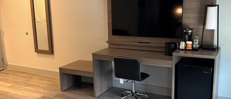A modern, compact living space with a wooden desk, a black office chair, a flat-screen TV, a small refrigerator, and a coffee maker.