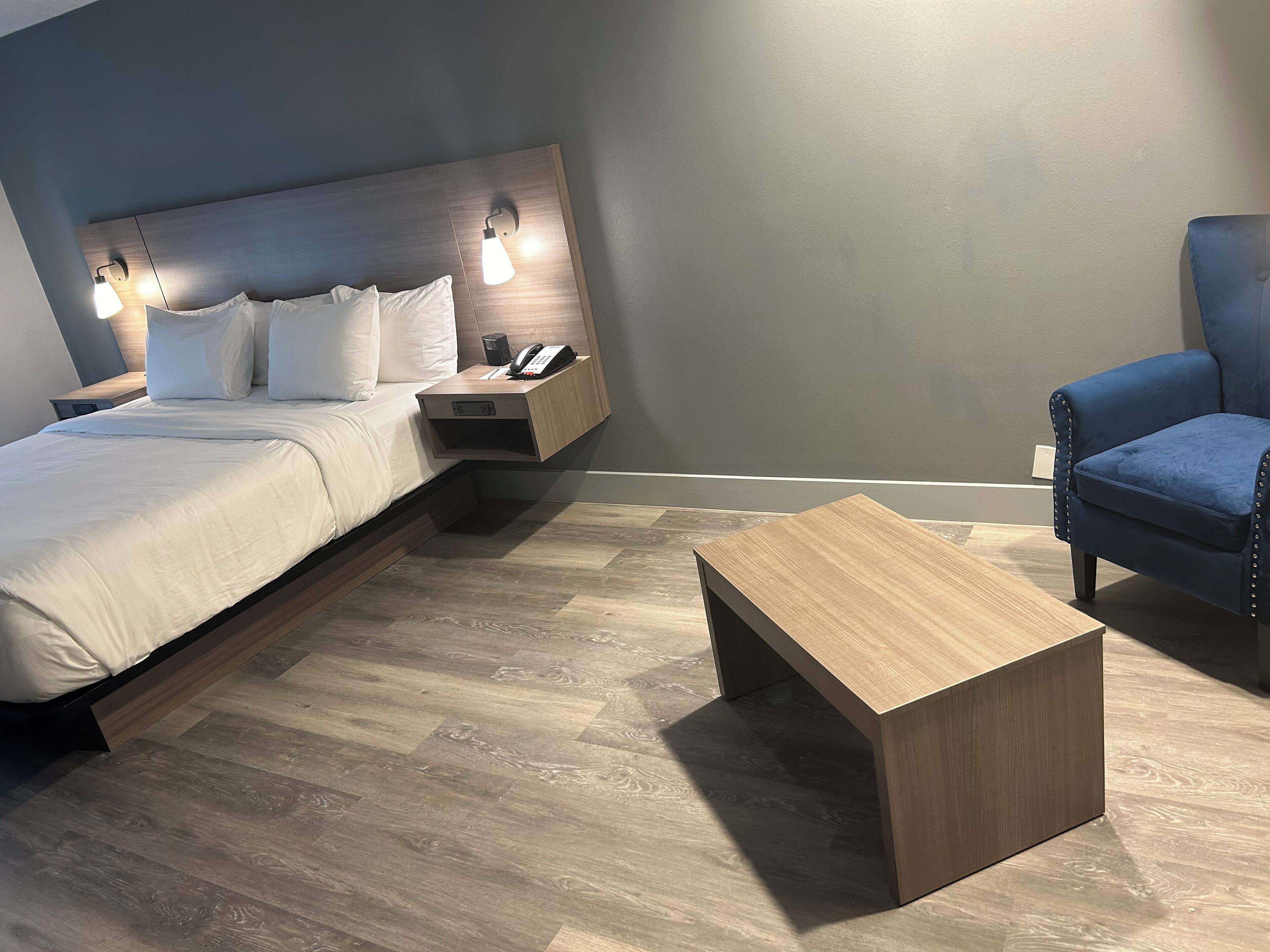 A modern hotel room with a bed, bedside lamps, a wooden nightstand, a blue armchair, and a wooden coffee table.