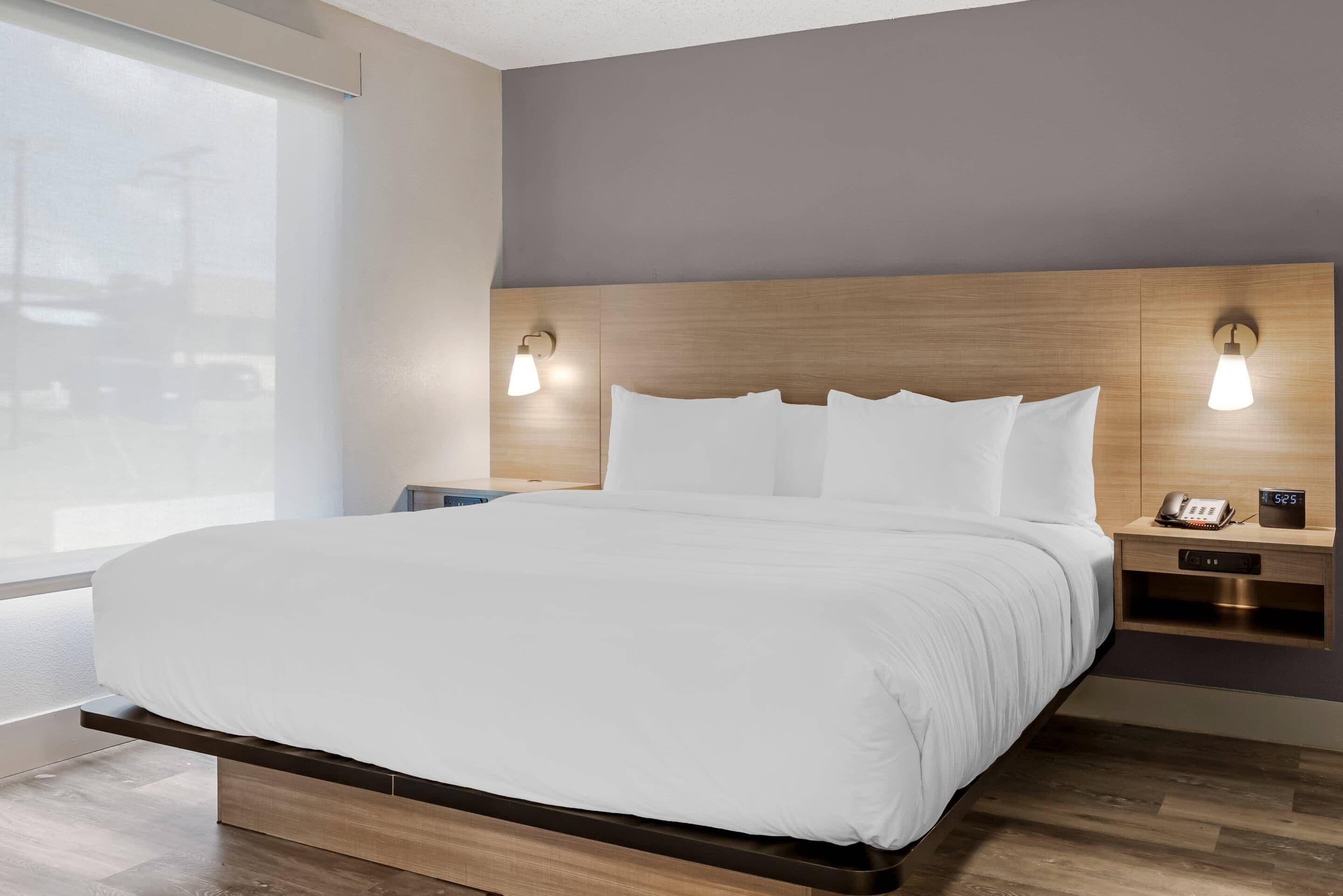 A hotel room with a large bed, wooden headboard, bedside lamps, a nightstand with a phone and clock, and a window with blinds.