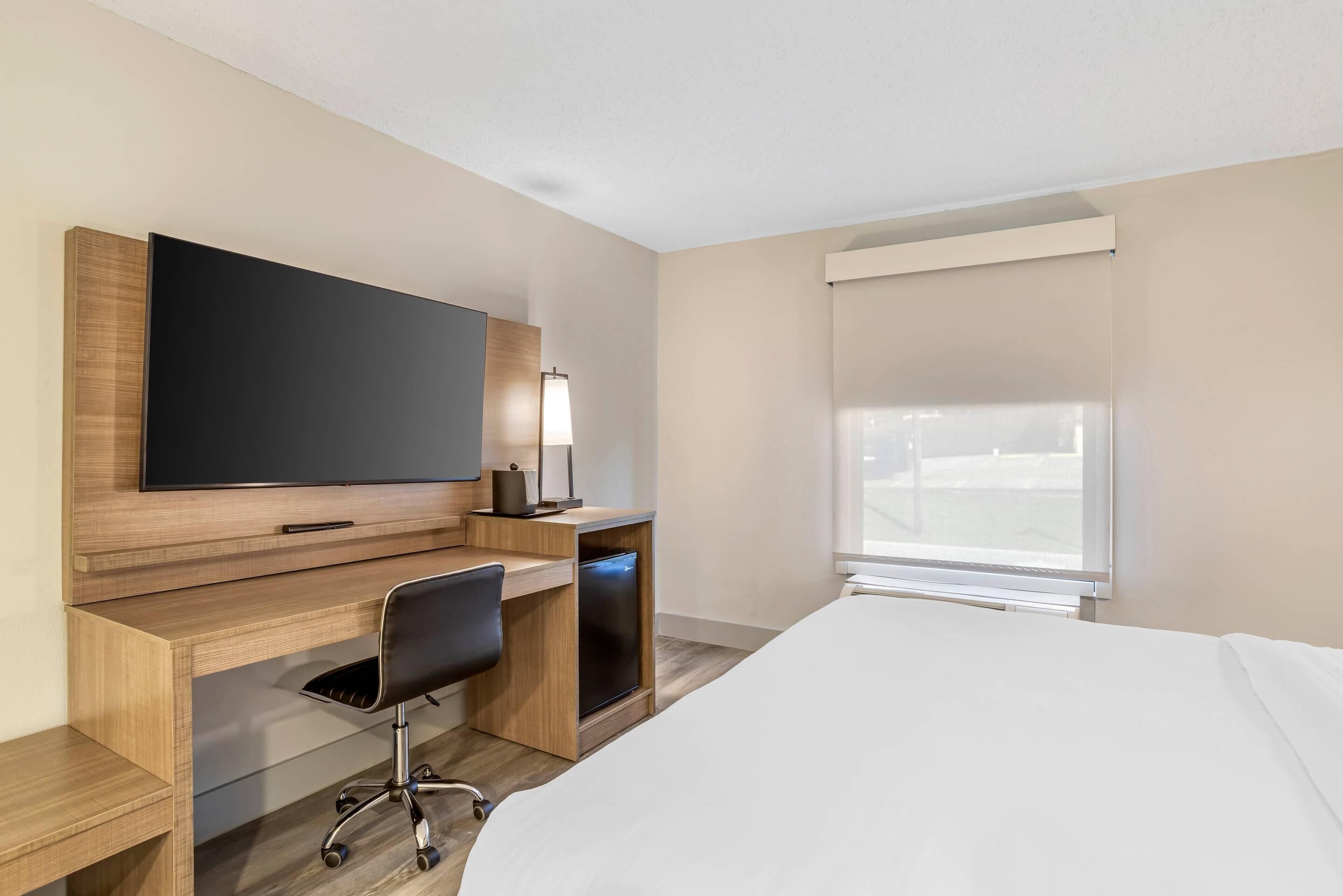 Een hotelkamer met een bed, een bureau met stoel, een televisie en een raam met jaloezieën.