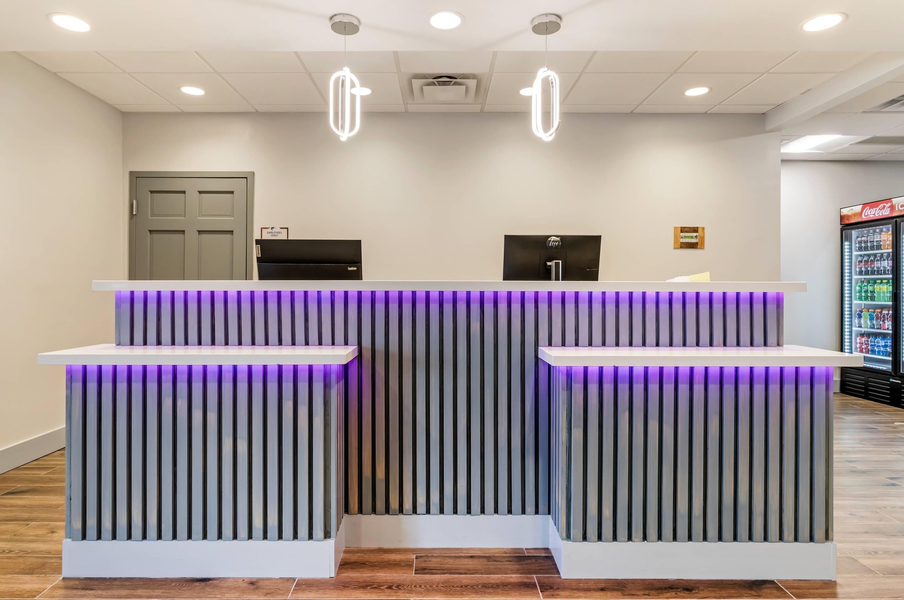 Un comptoir d’accueil au design moderne, deux écrans d’ordinateur, un réfrigérateur Coca-Cola et une porte visible au fond.