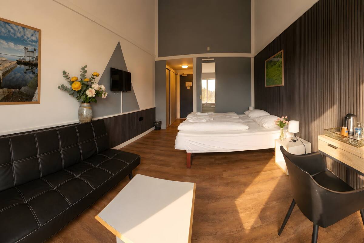 A hotel room with a bed, a black leather sofa, a white coffee table, a desk with a chair, a TV mounted on the wall, and a painting.