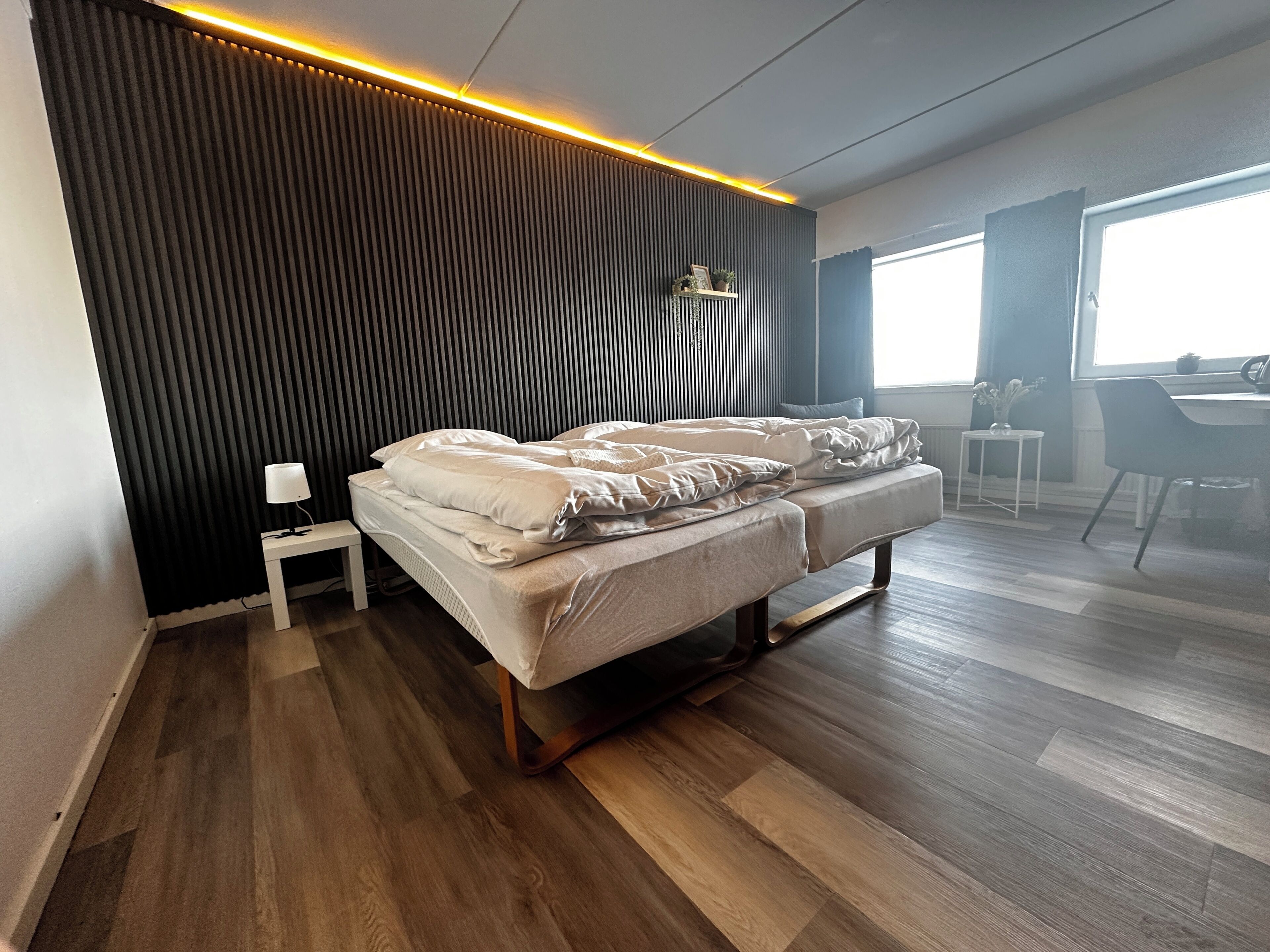 A modern bedroom with a bed, bedside table, lamp, and a view of the dining area.