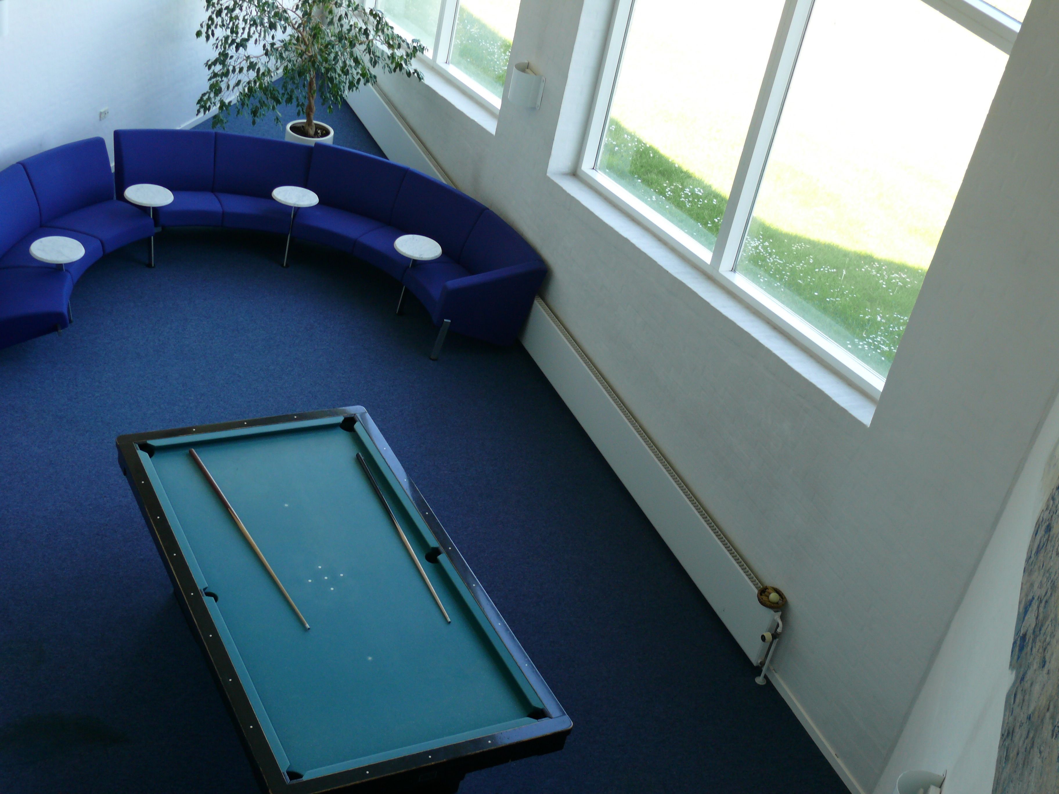 Una mesa de billar en una habitación con alfombra azul, un área de descanso azulada en forma de curva y un ventanal grande.