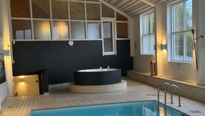 Ett modernt inomhuspoolområde med en stor rektangulär pool, en rund jacuzzi och en trappa som leder upp till en övre våning.