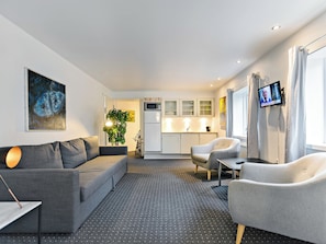 A modern living room with a grey sofa, a grey armchair, a small table, a potted plant, and a kitchen area with white cabinets and a microwave. - Løgstør Parkhotel (Logstor)