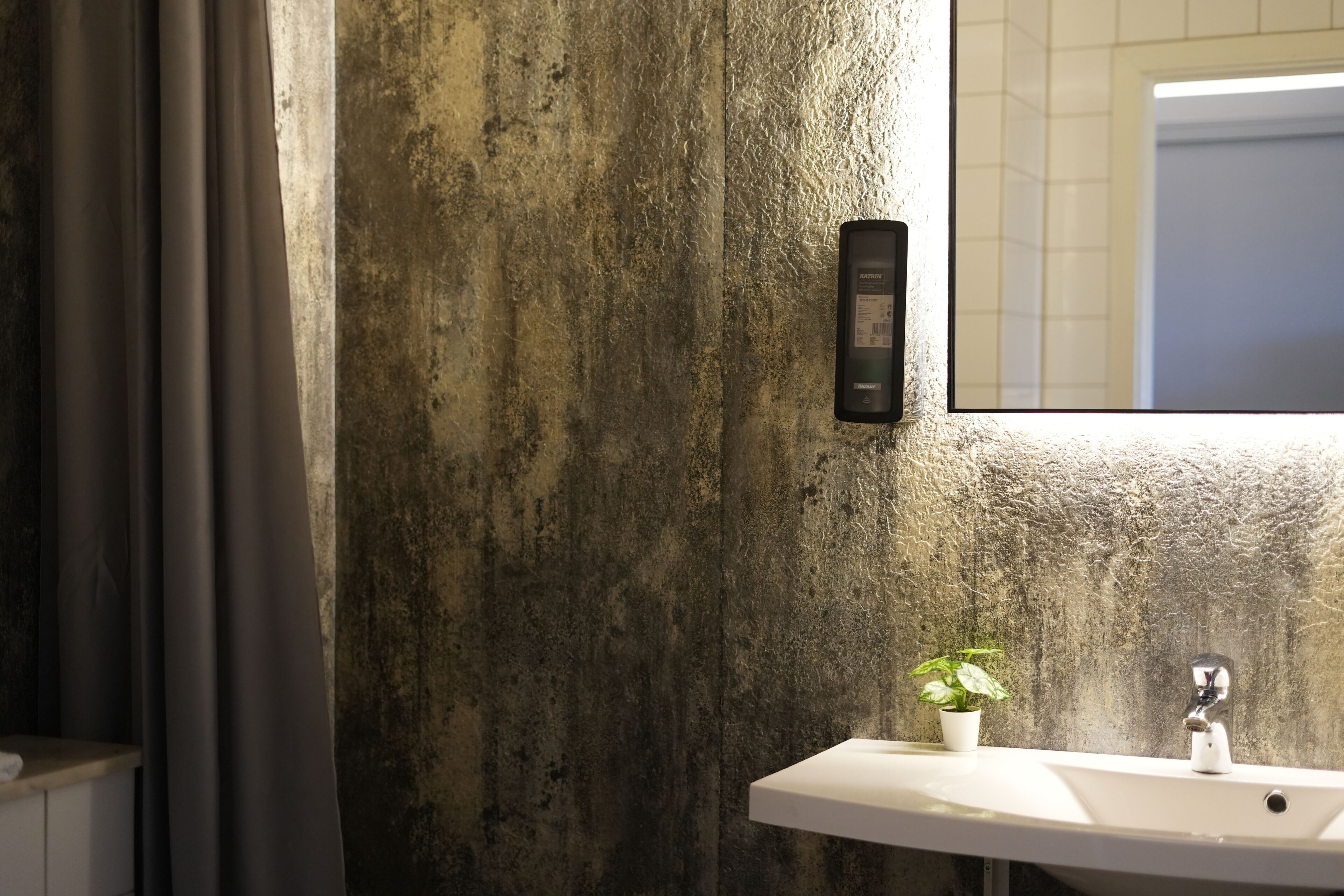 A bathroom with a concrete wall, a white sink, a small potted plant, and a wall-mounted light fixture.
