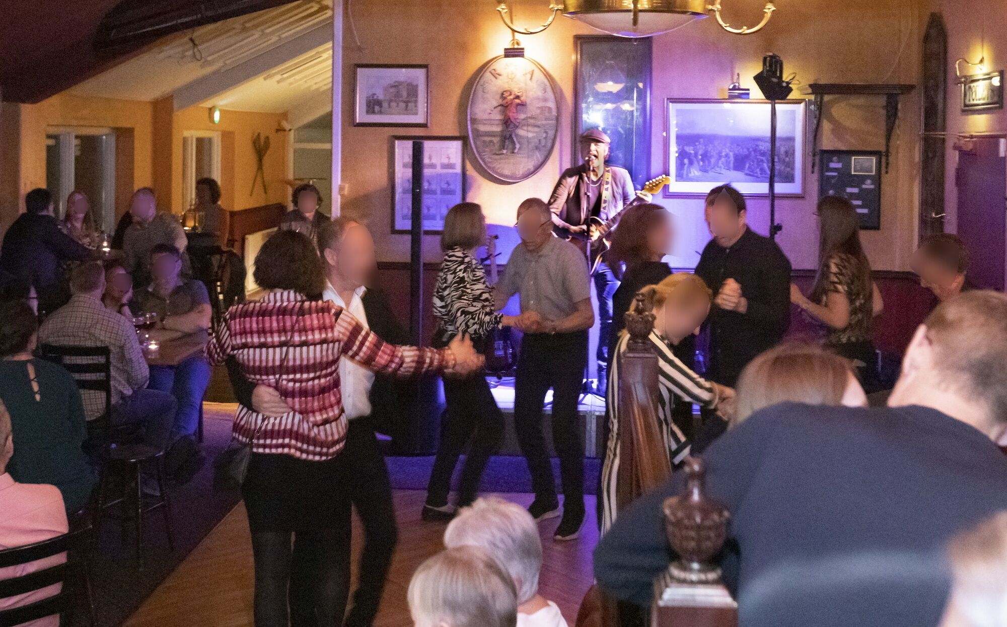 A group of people dancing in a dimly lit room with a stage, tables, and chairs.