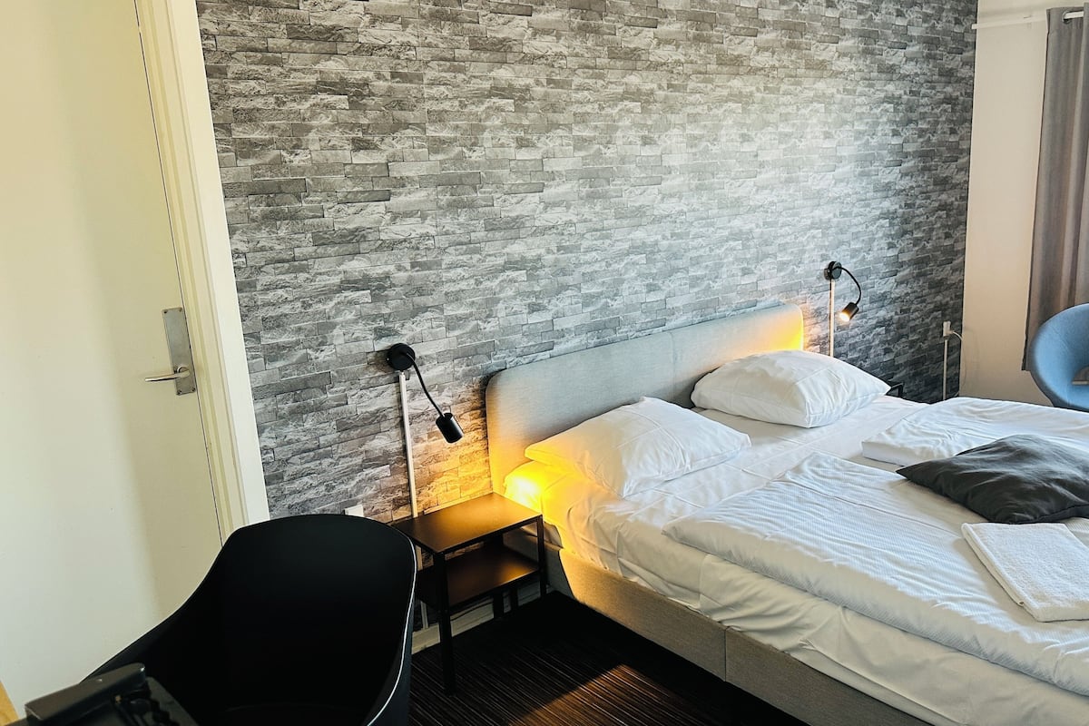 A hotel room with a stone-textured wall, a bed with white bedding, a desk with a chair, and a small table with a lamp.