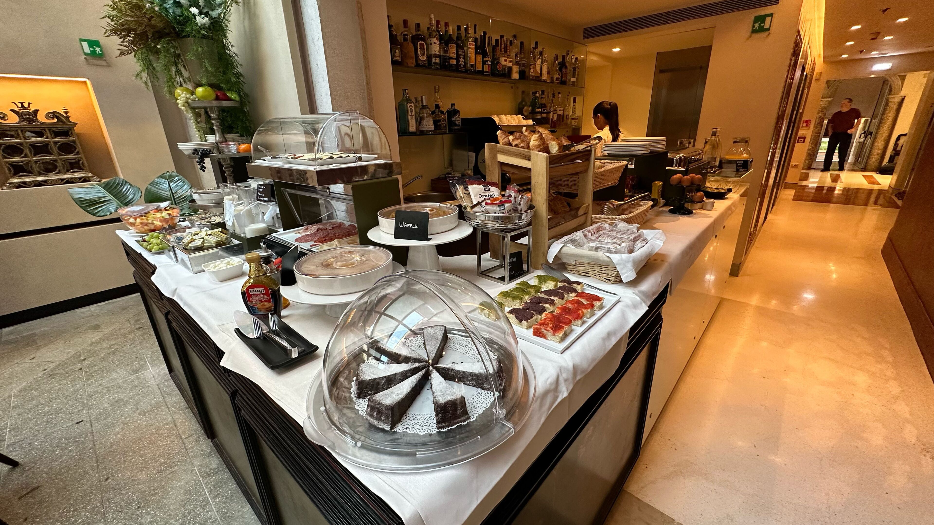 A buffet area with various food items, including a cake under a glass dome, a waffle station, and a selection of cheeses and meats.