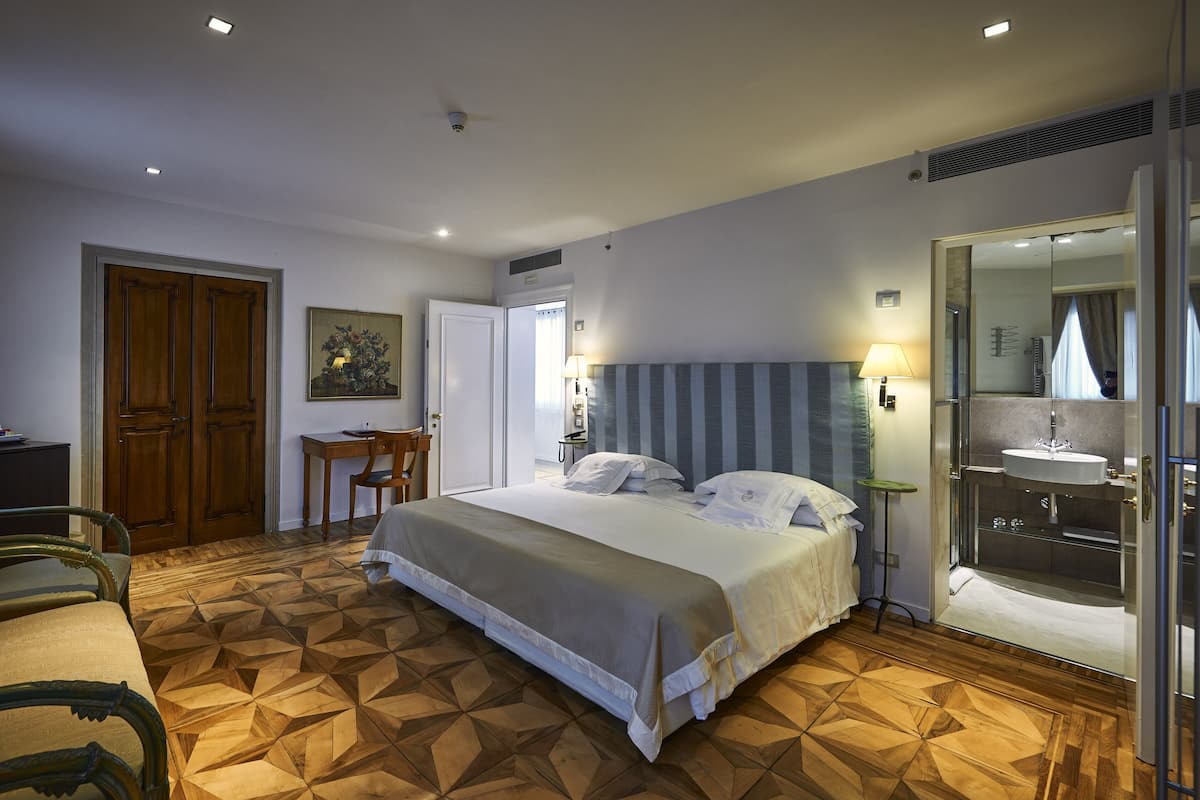 A hotel room with a bed, a desk, a chair, a painting, and a bathroom with a sink and mirror.