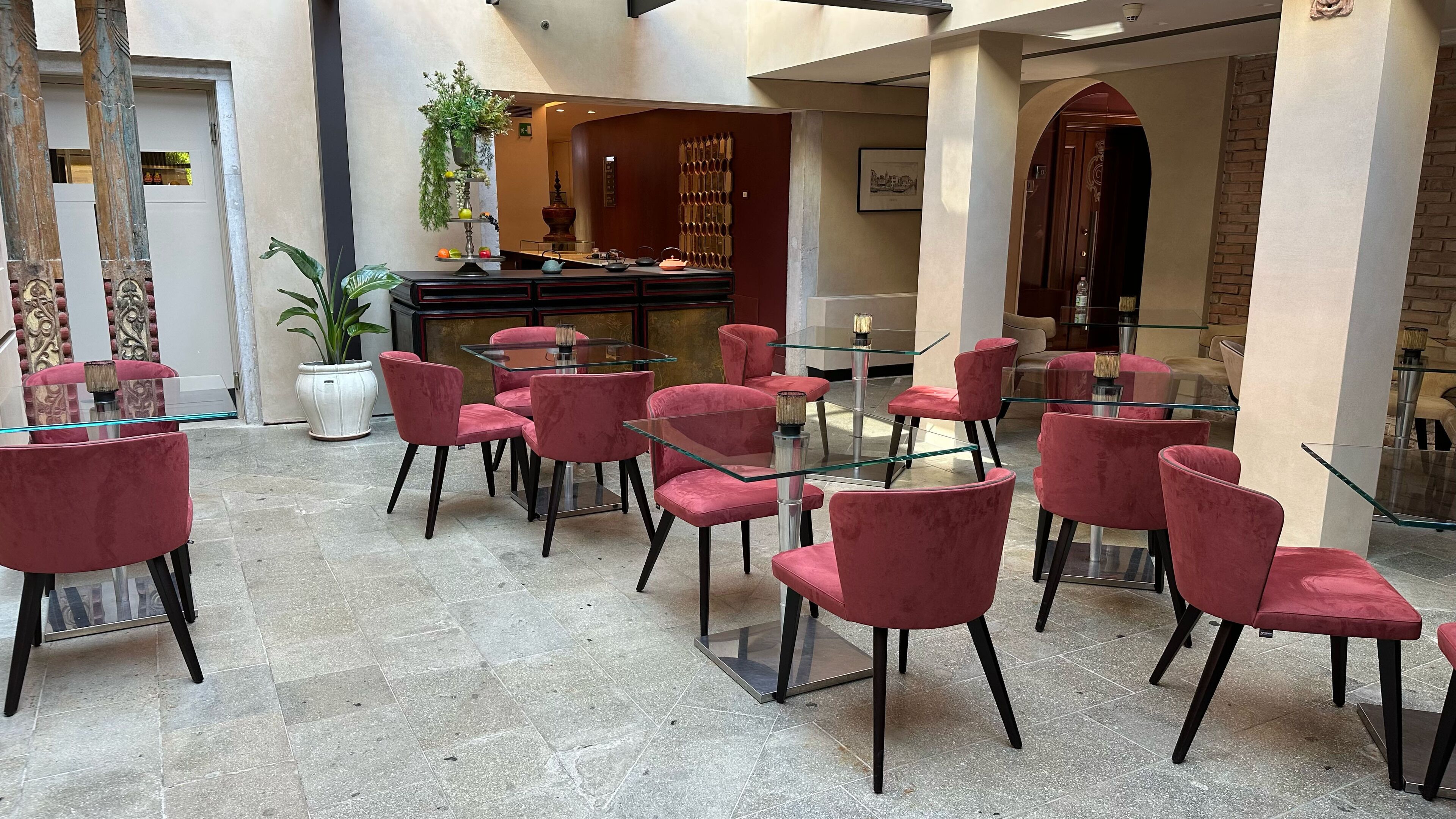 A hotel lobby with red velvet chairs and glass tables, a potted plant, and a bar area.