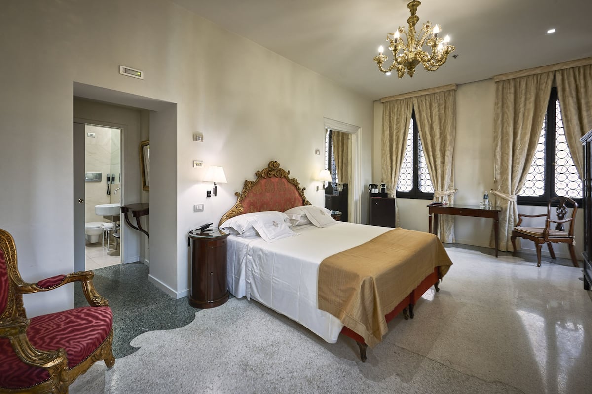 A hotel room with a large bed, a sitting area with a chair, a desk, and a chandelier.