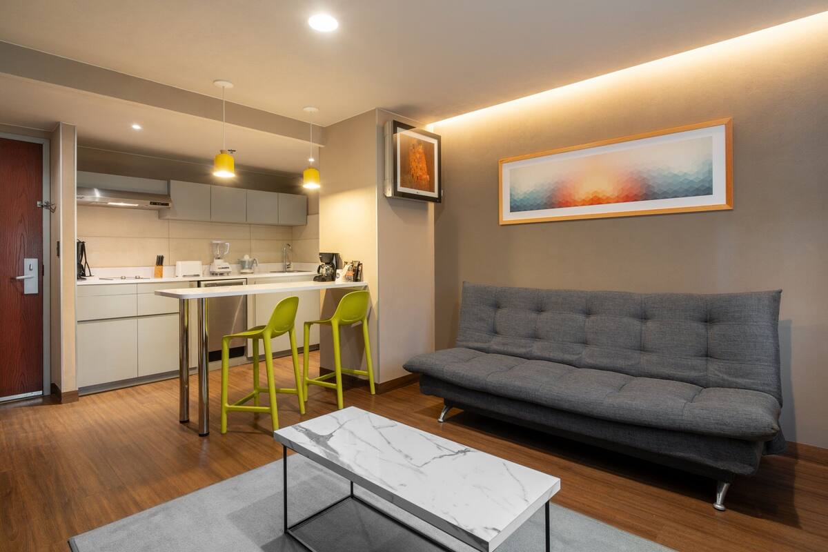 Una sala de estar moderna con un sofá gris, una mesa de centro blanca y una cocina con una mesa de comedor y sillas.