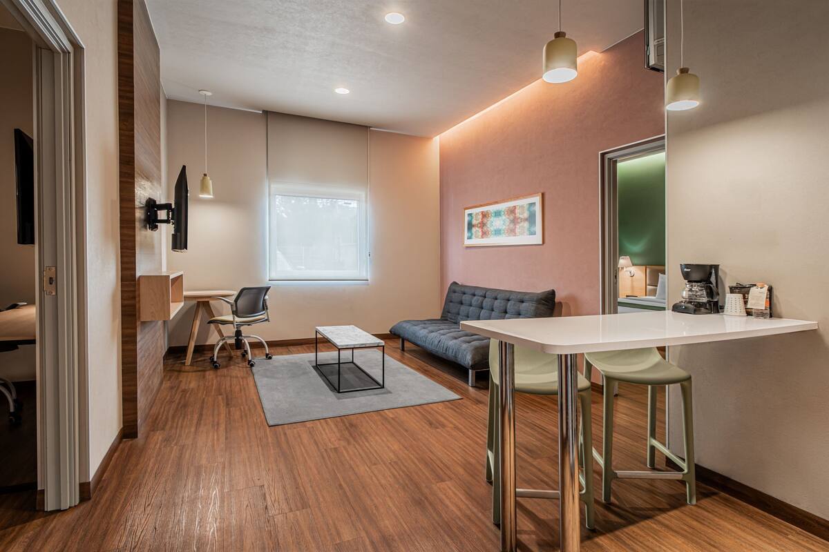 Una habitación moderna y bien iluminada con suelo de madera, un escritorio blanco, un sofá gris y una pequeña mesa.