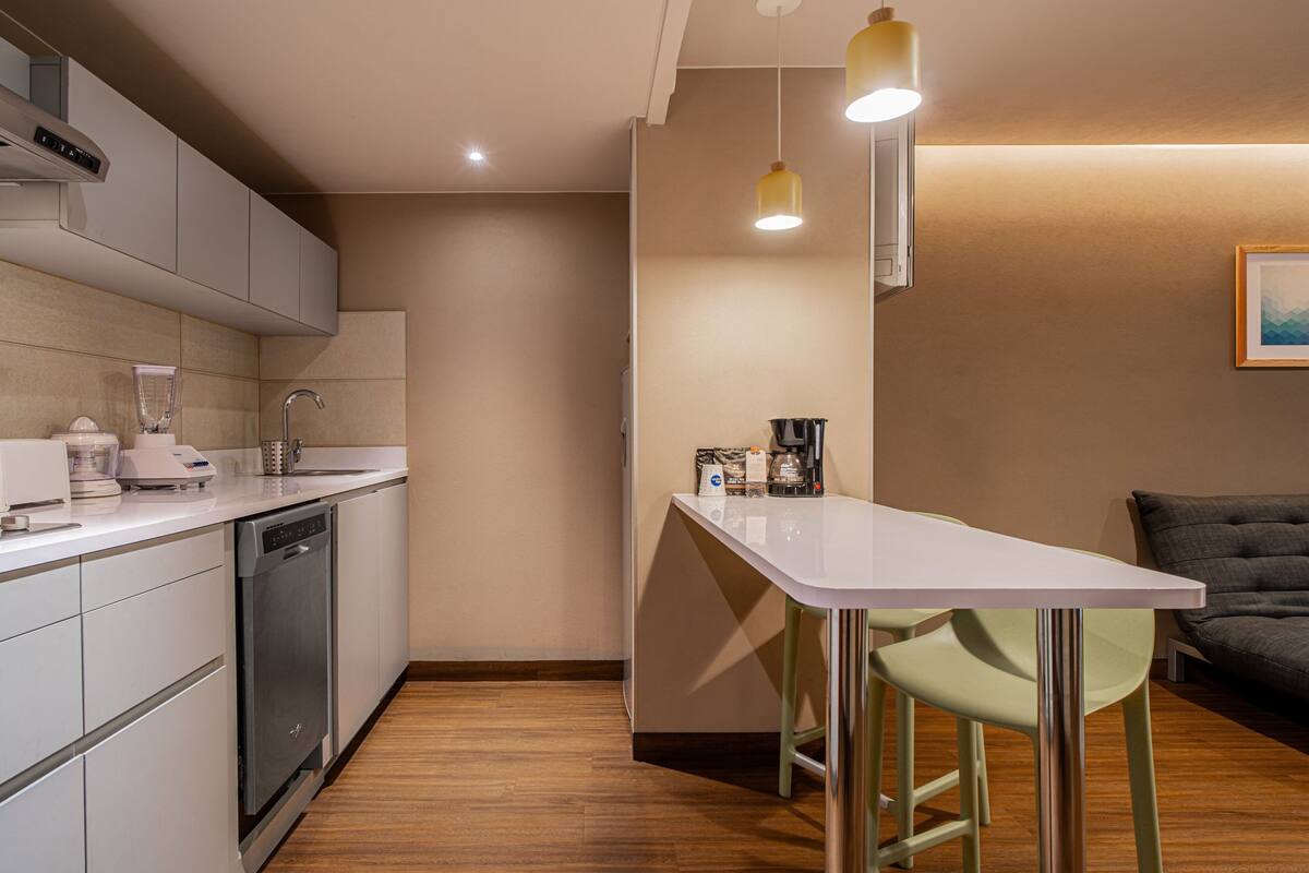 Una cocina moderna con armarios blancos, lavavajillas, cafetera y un taburete de bar.