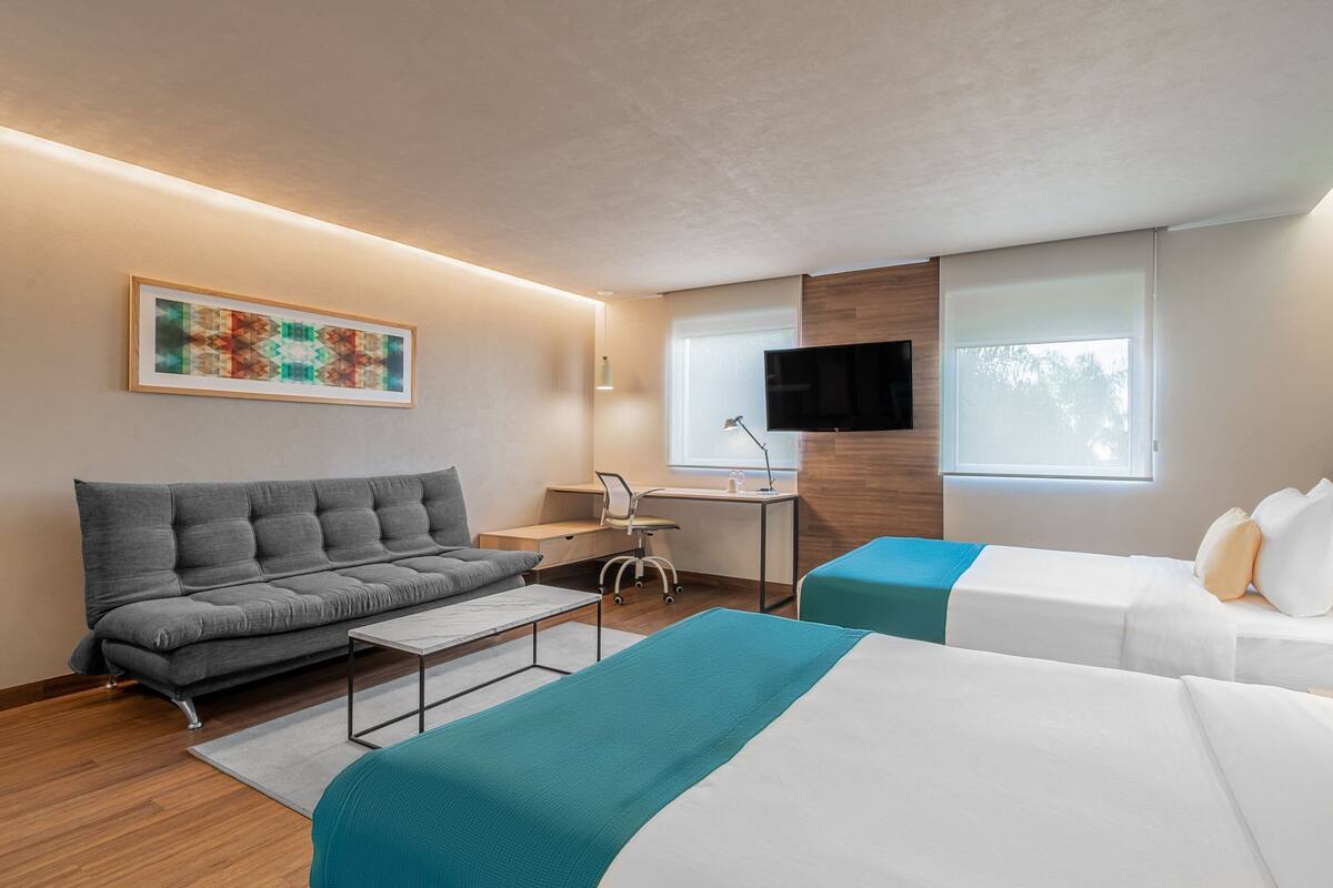 Una habitación de hotel moderna con sofá, dos camas, un escritorio, televisor y ventana.