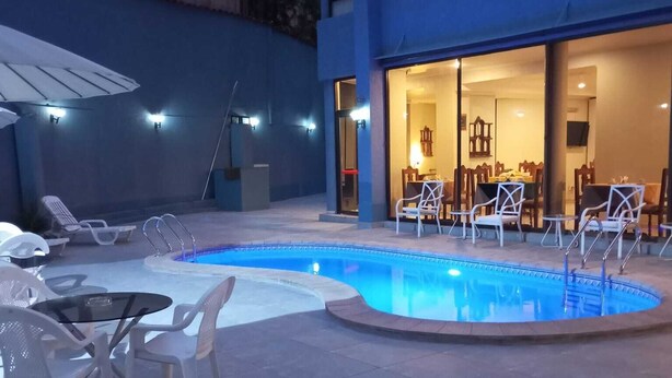 A swimming pool with lounge chairs and tables, a covered seating area, and a building entrance with a dining area visible inside. - Hotel Alcala del Rio (Santiago)