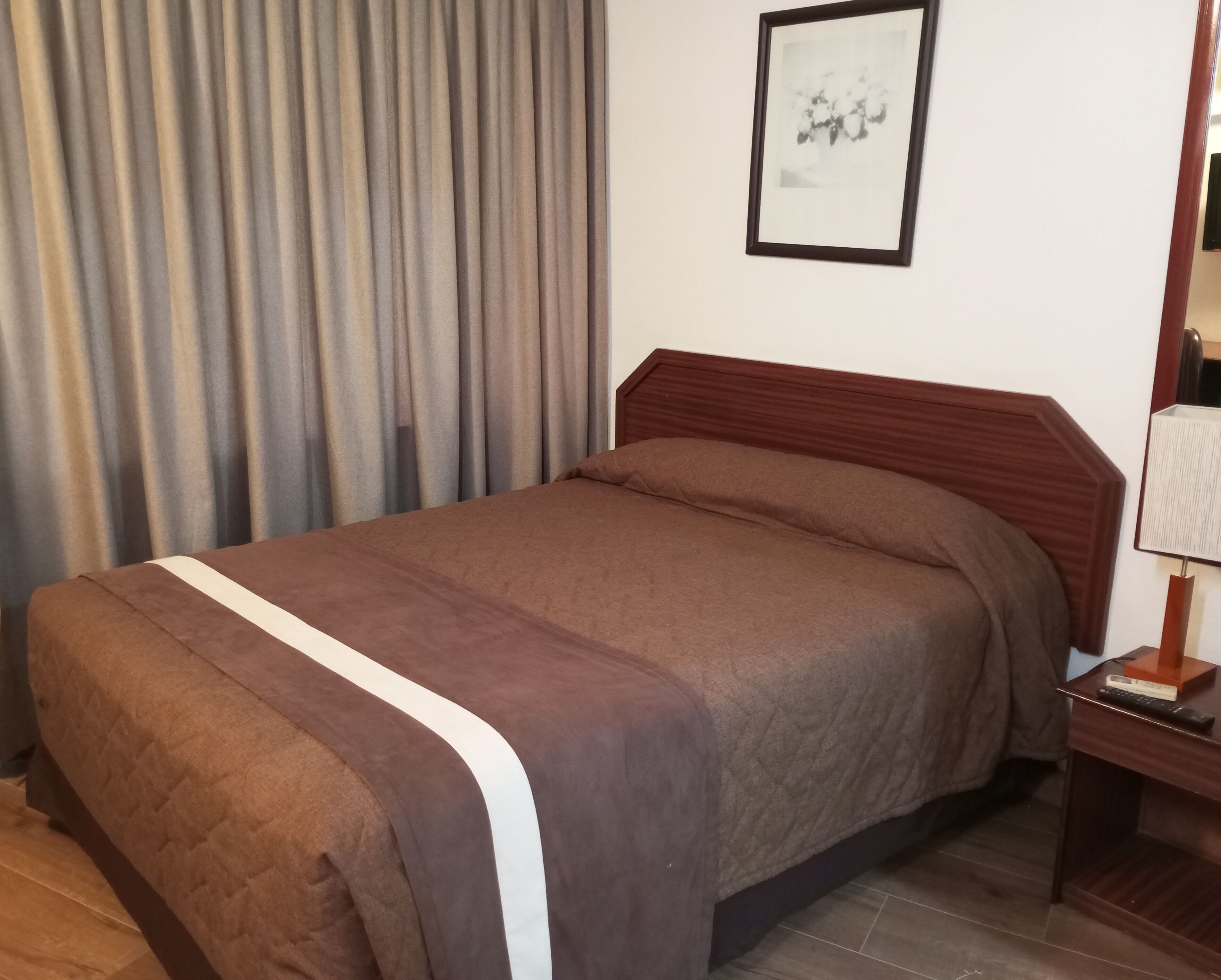 A neatly made bed with a brown bedspread and a white stripe, a wooden headboard, a framed picture on the wall, and a bedside table with a lamp and a remote control.