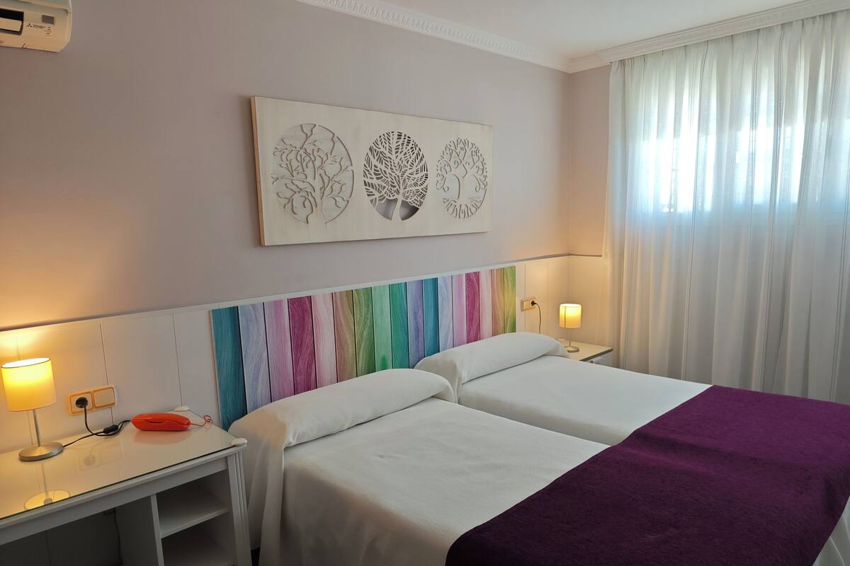 Une chambre d’hôtel avec deux lits, un bureau avec une lampe, une œuvre d’art accrochée au mur et une fenêtre avec des rideaux.