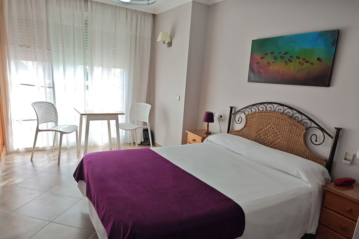 Une chambre à coucher avec un lit, une chaise, une table, une lampe et un tableau accroché au mur.