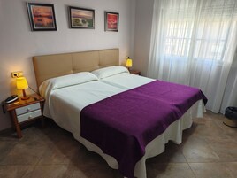Una cama bien hecha con una colcha morada, una lámpara de noche y una mesita con un teléfono.