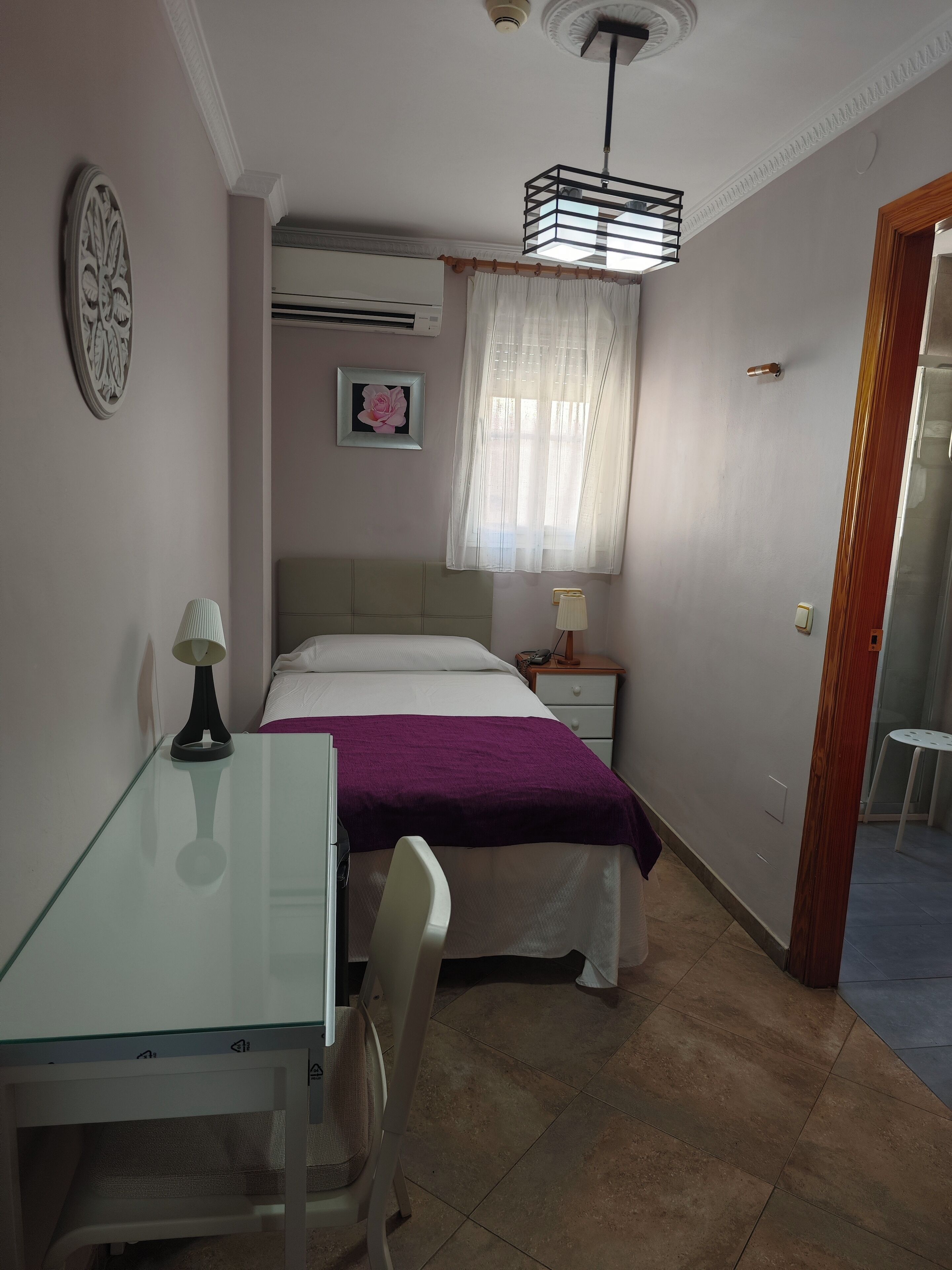 A bedroom with a bed, a desk, a chair, a bedside table with a lamp, a window with sheer curtains, and a ceiling fan.