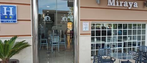 The exterior of Hotel Miraya with a glass entrance, outdoor seating area, and a sign indicating a local climate zone.