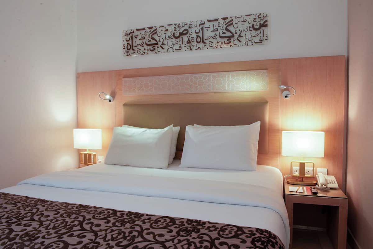 Una habitación de hotel con una cama, dos lámparas de noche, una mesita de noche con un teléfono y un adorno en la pared.