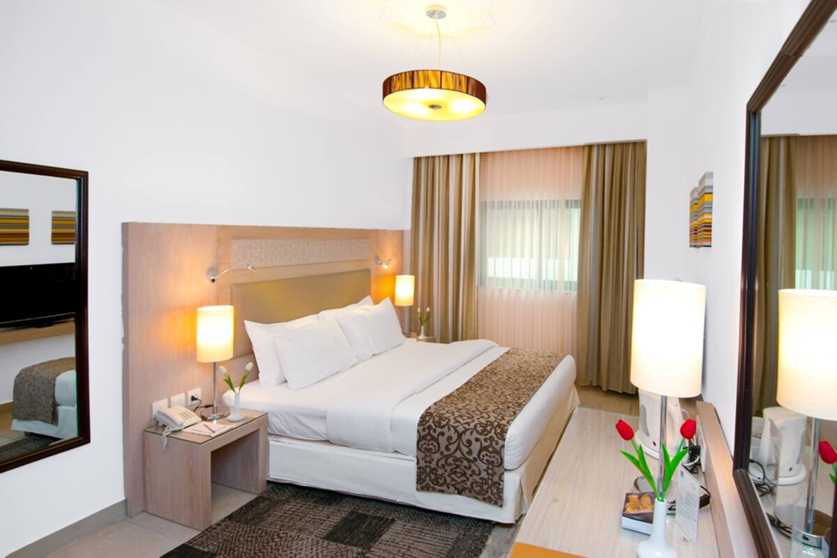 Een hotelkamer met een bed, nachtlampjes, een nachtkastje met een telefoon, een spiegel en een raam met gordijnen.