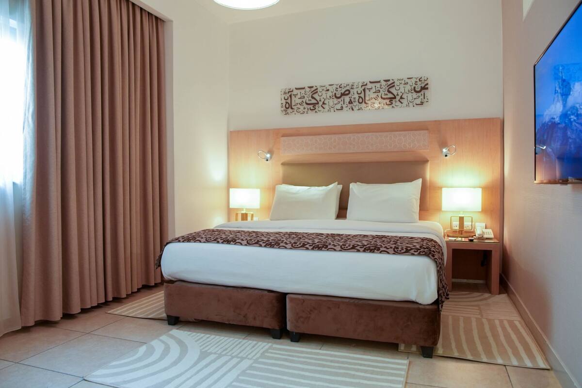 Een hotelkamer met een groot bed, twee nachtkastjes met lampen, een hoofdbord met decoratief paneel, een groot raam met gordijnen, en een kunstwerk aan de muur.