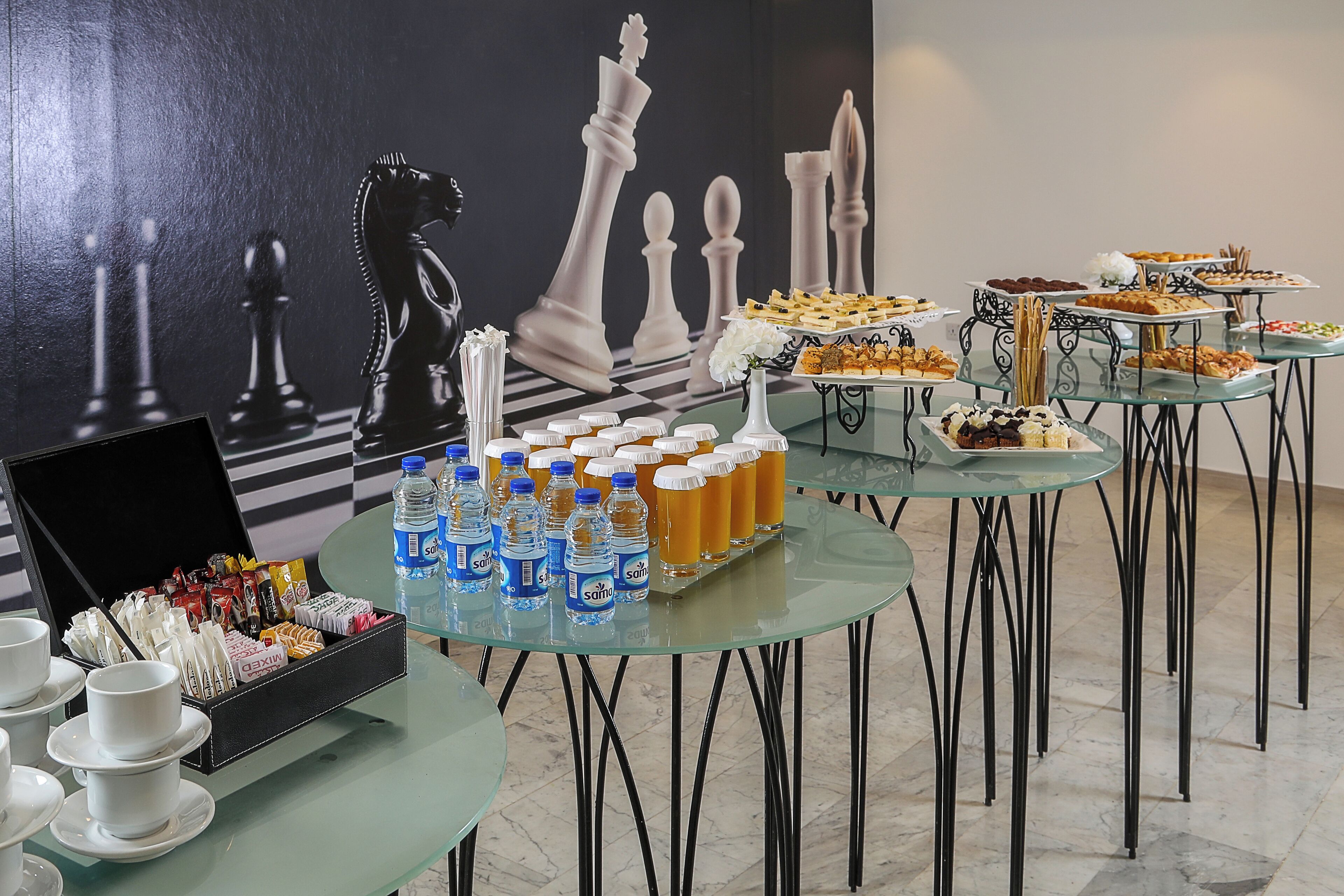 A buffet setup with various food items and beverages on glass tables, with chess pieces in the background.