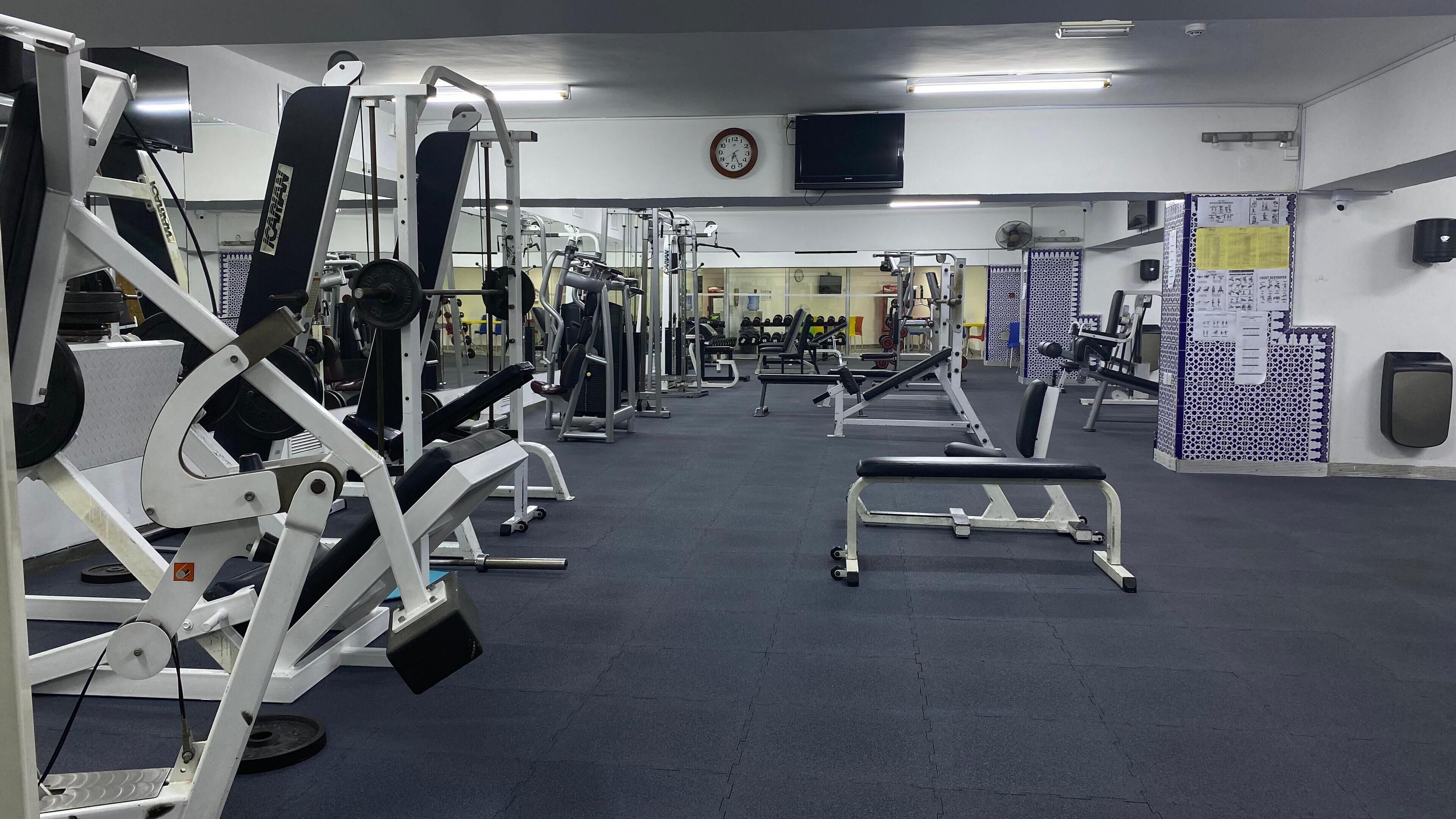 Ein Fitnessraum mit unterschiedlichen Trainingsgeräten, einer Uhr an der Wand und einem Fernseher über einer Theke.