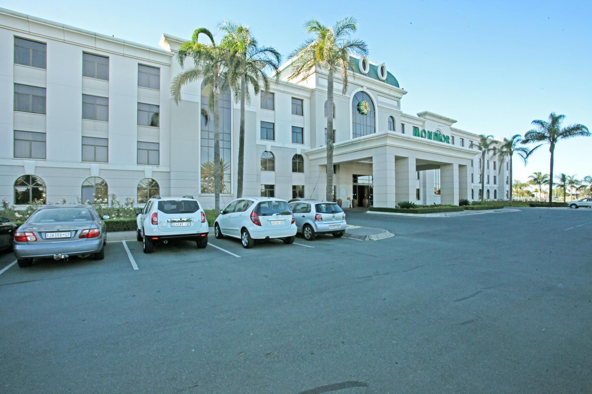 A hotel with a white facade, palm trees, and a parking lot with several cars.