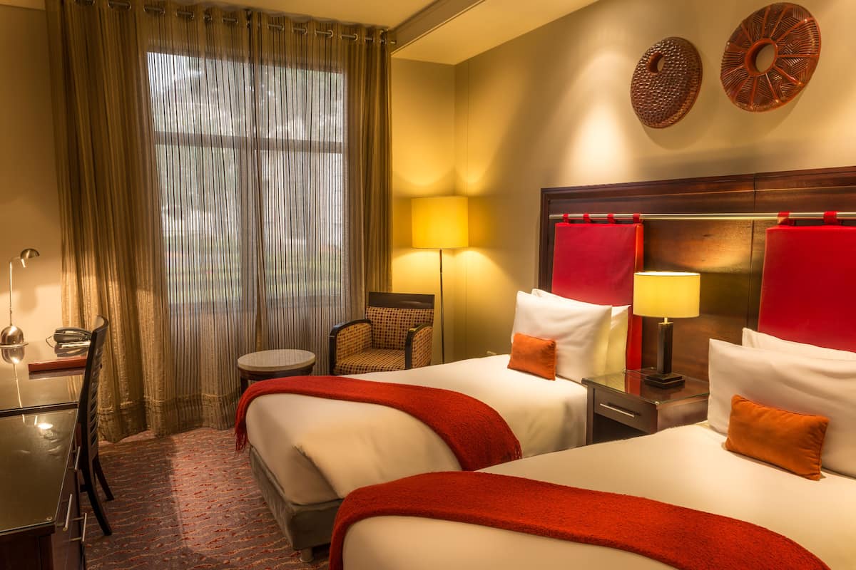 Une chambre d’hôtel avec deux lits, un bureau, une chaise, une lampe et une fenêtre avec des rideaux.