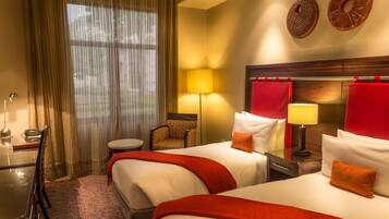 A hotel room with two beds, a desk, a chair, a lamp, and a window with curtains.