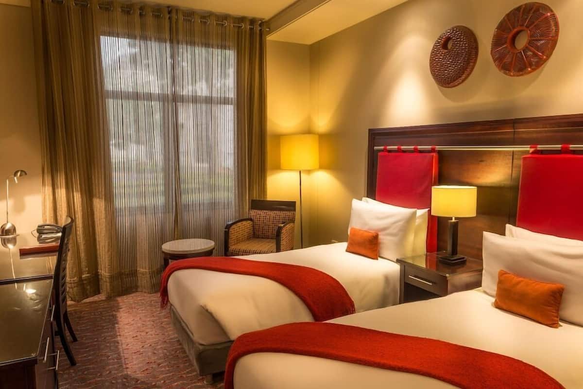 Une chambre d’hôtel avec deux lits, un bureau, une chaise, une lampe et une vue par une fenêtre avec des rideaux.