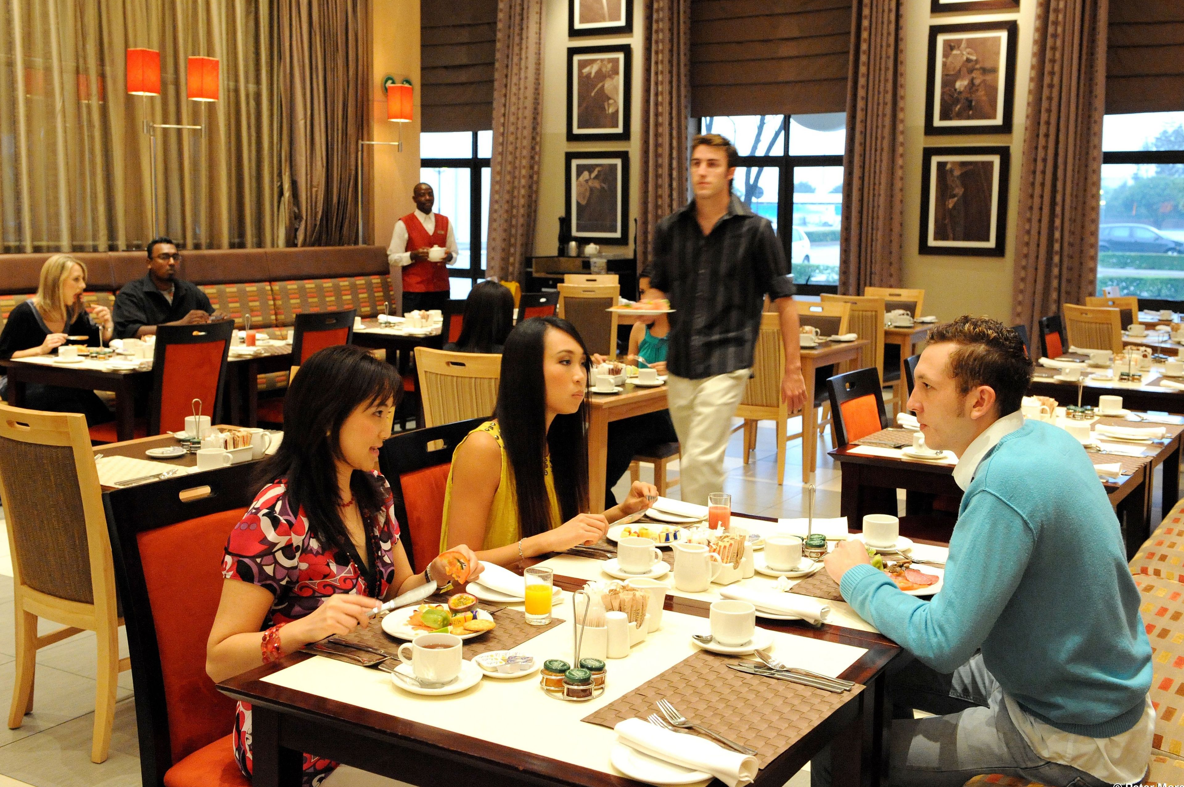 A restaurant with multiple tables and chairs, diners seated, and a waiter attending to guests.