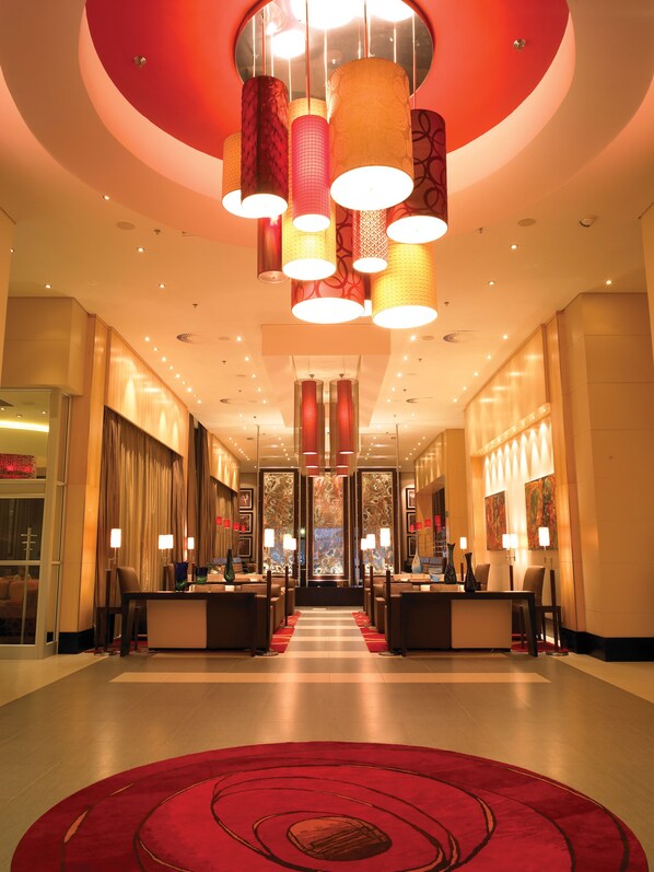 A hotel lobby with a central reception desk, multiple seating areas, and a red carpet leading to the entrance.