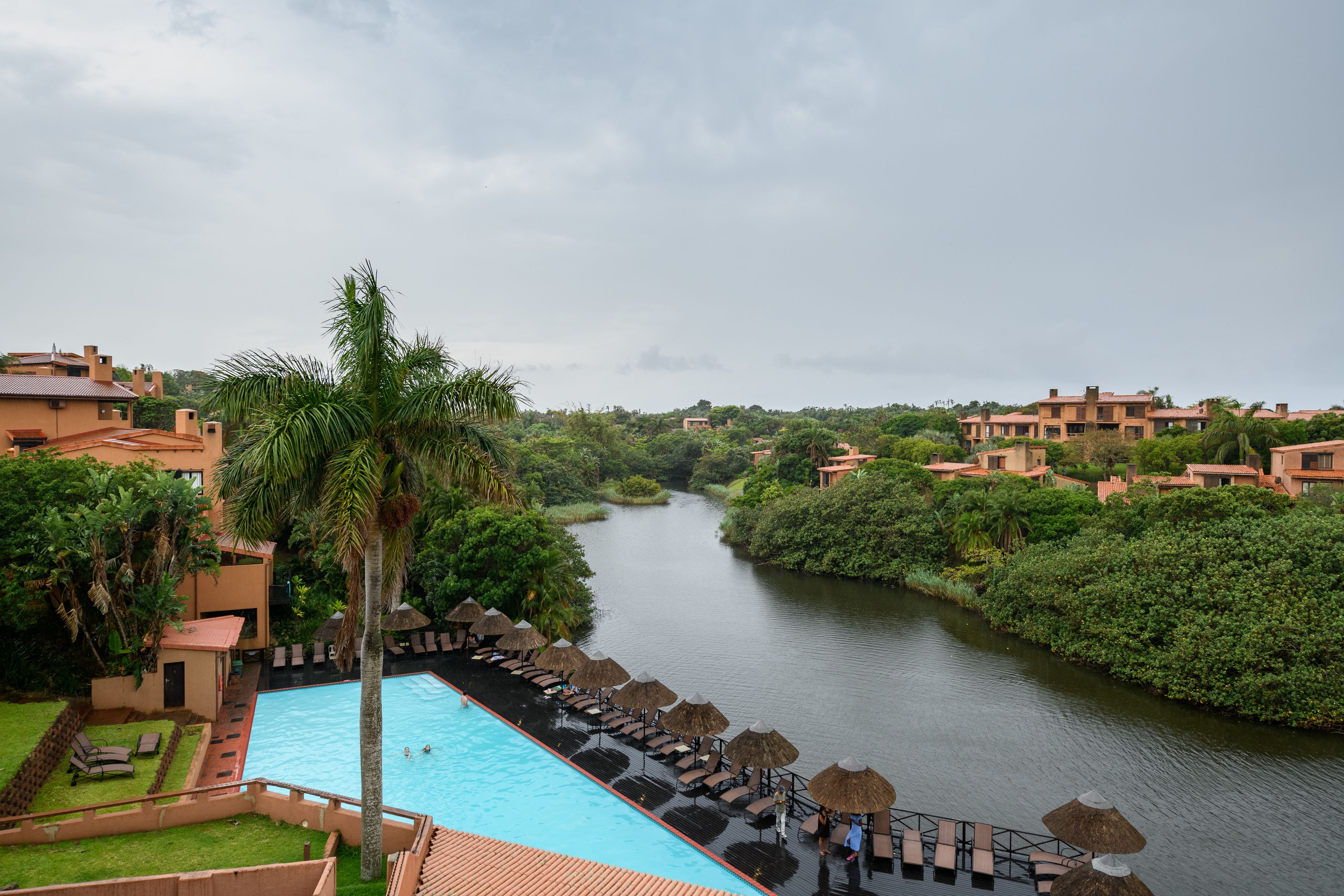 A pool area with lounge chairs and umbrellas, surrounded by lush greenery and a river.