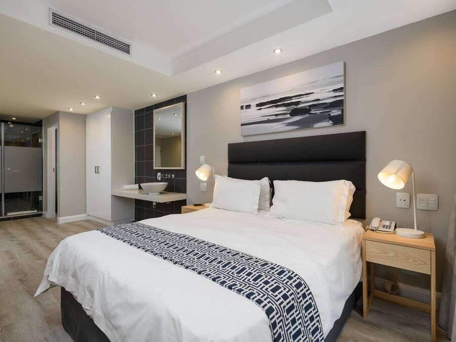 Quarto de hotel moderno com uma cama grande, luminárias de cabeceira, mesa de cabeceira, banheiro com pia e um espelho amplo.