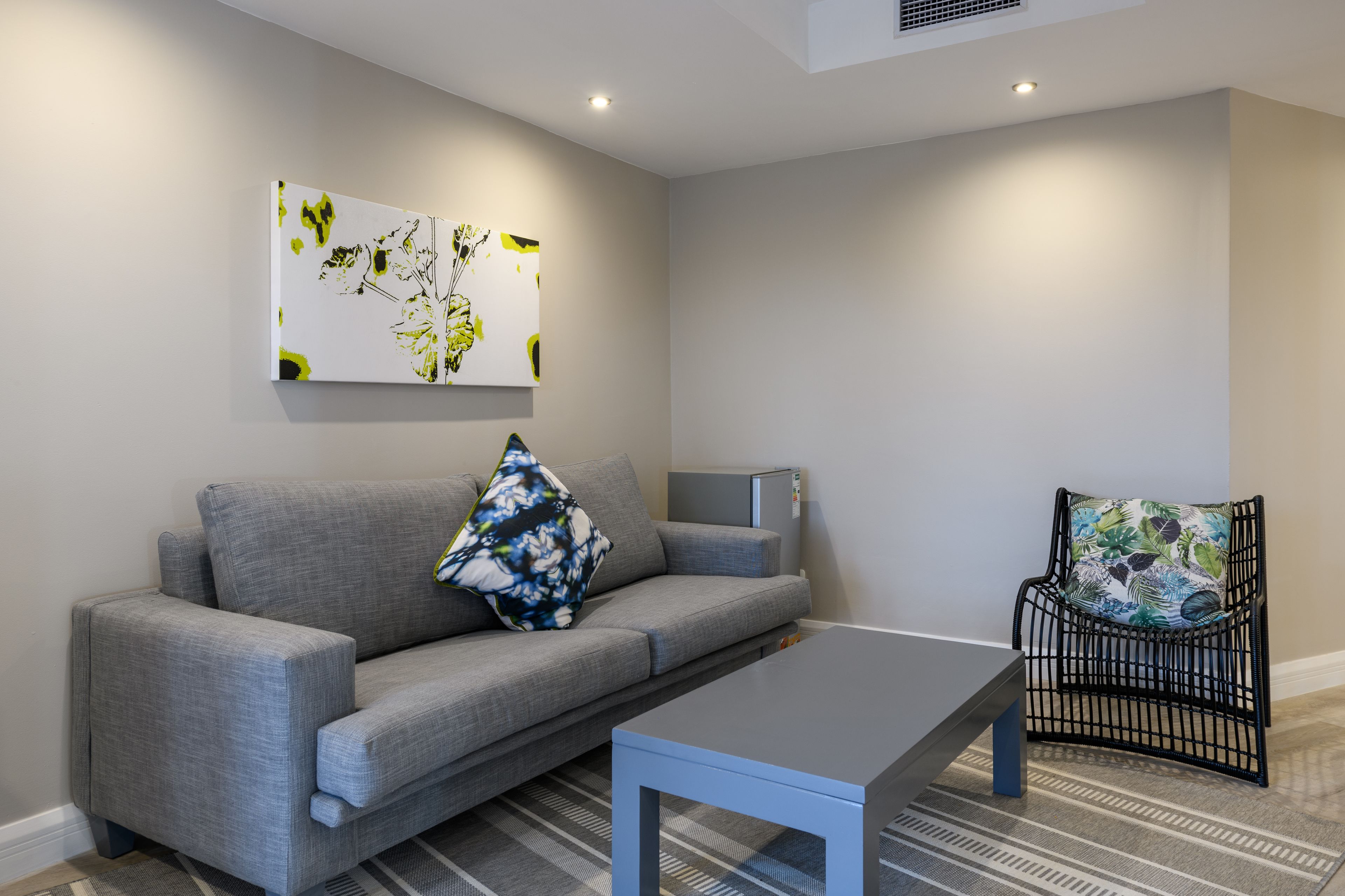 A modern living room with a grey sofa, a coffee table, a black wicker chair, and a wall-mounted flat-screen TV.