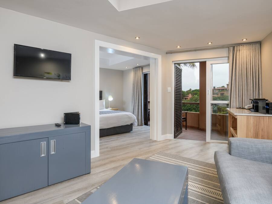A modern hotel room with a flat-screen TV, a grey sofa, a wooden desk, and a balcony with a view.