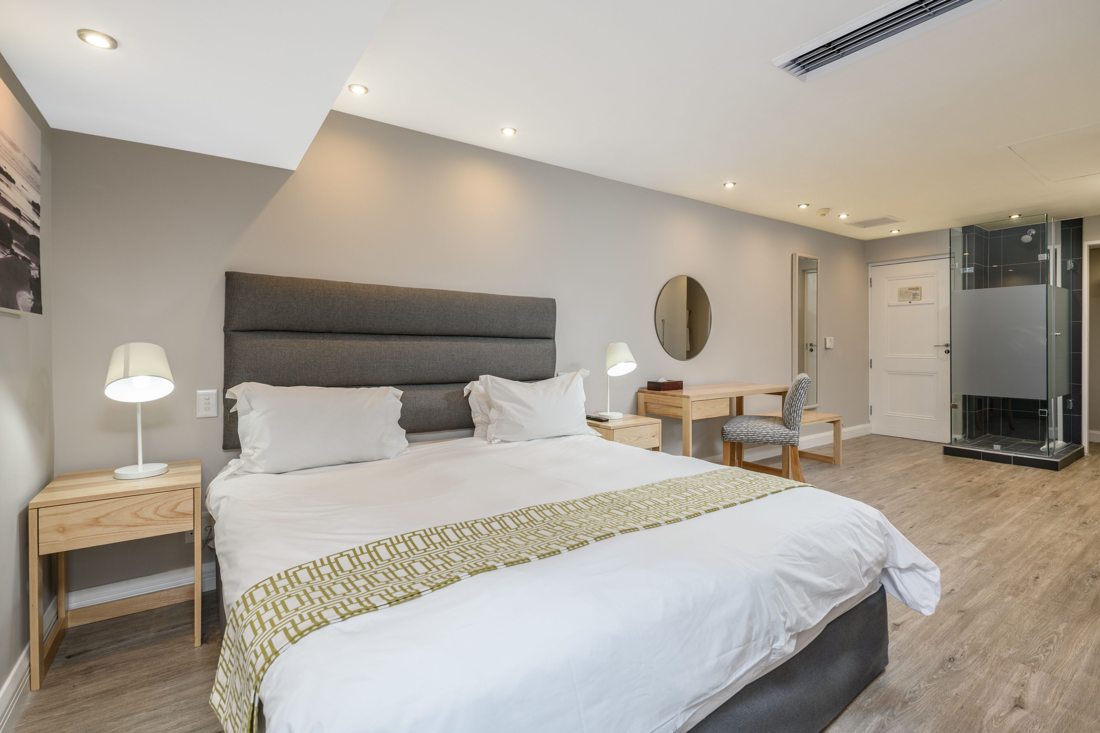 A modern hotel room with a large bed, bedside tables, a desk, and a glass shower enclosure.
