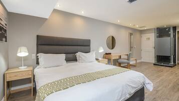 A modern hotel room with a large bed, bedside tables, a desk, and a glass shower enclosure.