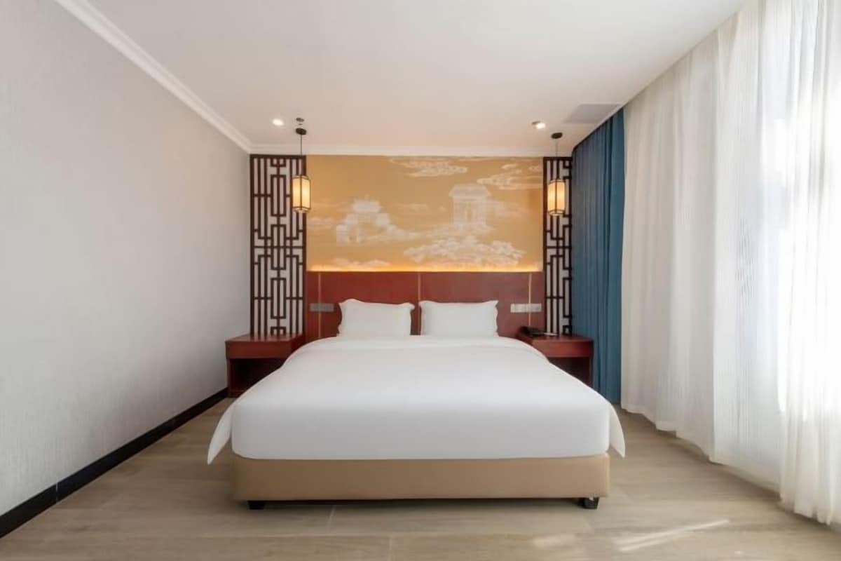 A hotel room with a large bed, a headboard with a decorative panel, two bedside tables with lamps, and a large window with curtains.
